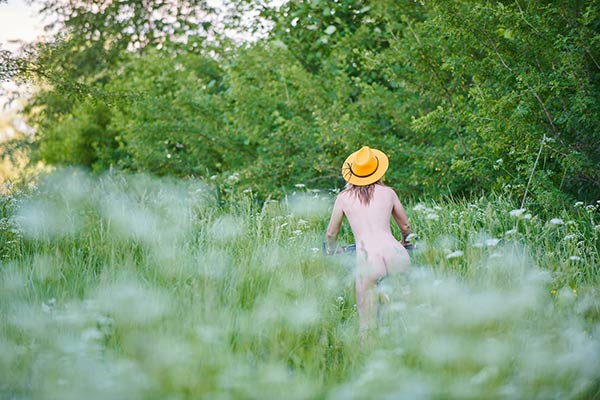 Blumenwiese - man sieht von hinten eine nackte Radfahrerin mit gelbem Hut